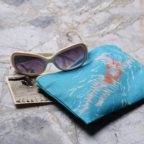 Mise en scène d'une pochette à zip en tissu avec une paire de lunettes de soleil et une photographie argentique noir et blanc. Le motif sur la pochette représente une nageuse avec un maillot de bain orange sur fond bleu