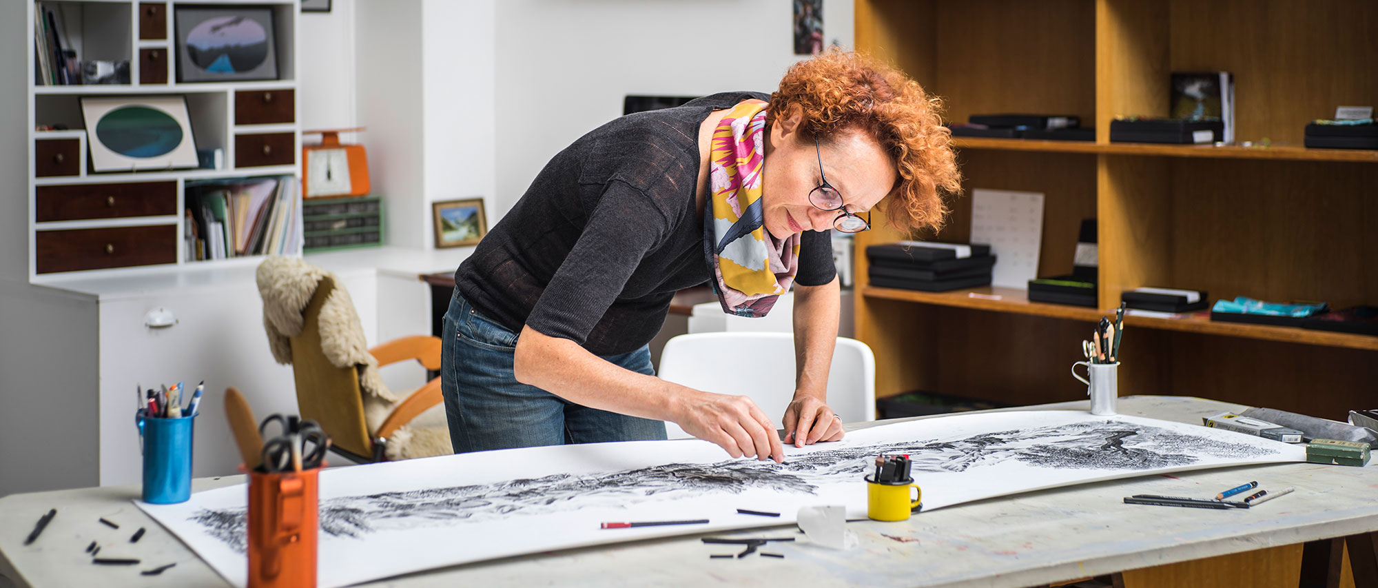 Dans un atelier d'artiste, Céline Dominiak est debout, en train de dessiner, penchée sur un grand dessin au fusain. elle porte un foulard.